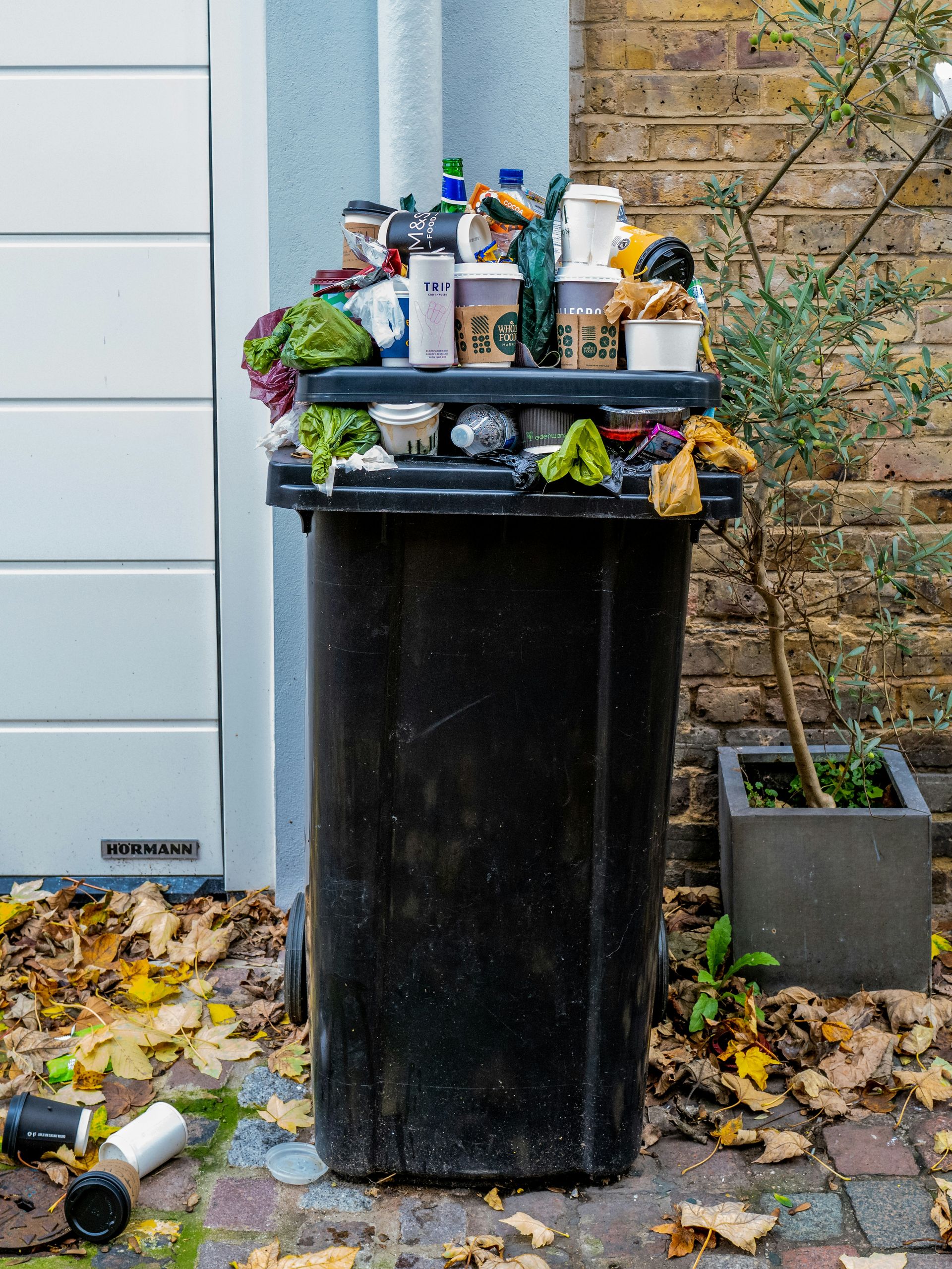 An overflowing black plastic trash bin filled with coffee cups and plastic bags